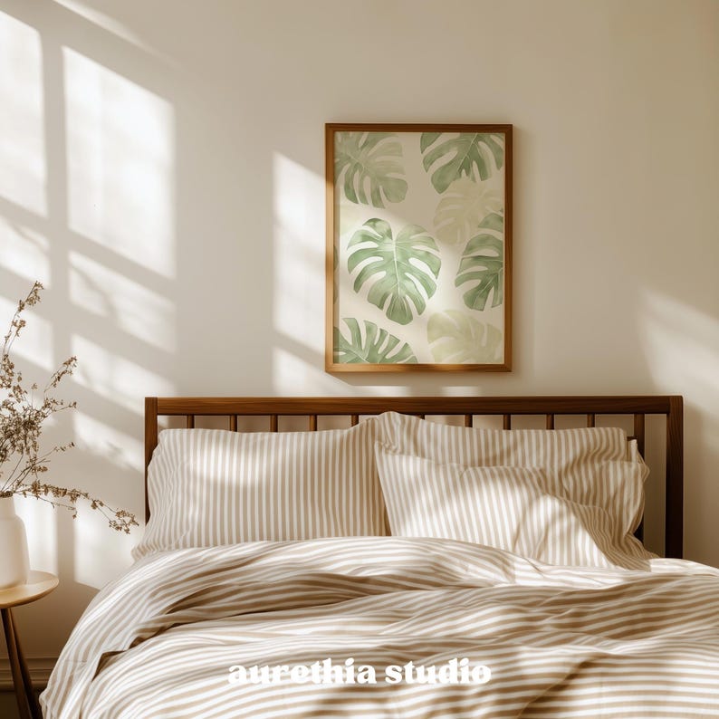 May include: A bedroom scene featuring a bed with striped bedding and a wooden headboard. Above the bed, a framed print displays watercolor-style monstera leaves in shades of green. The room is bathed in natural light, creating a warm and inviting atmosphere.