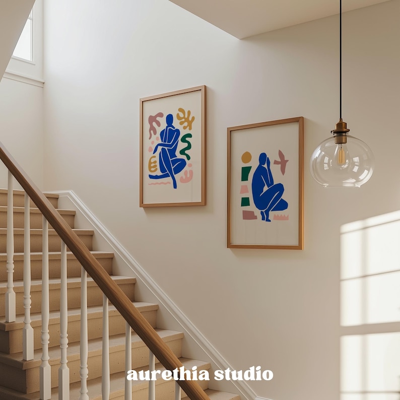 May include: Two framed art prints with abstract figures in blue, yellow, pink, and green hang on a white wall above a wooden staircase. The prints are in wooden frames. The text "aurethia studio" is at the bottom.