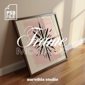 May include: A framed art print mockup leaning against a wall. The artwork features a black starburst design on a pink background, with the words "Frame Mockup" in white script. The frame is a dark brown color.