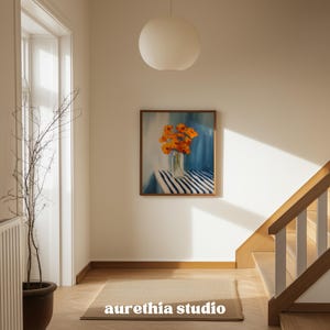 May include: A framed painting depicts orange flowers in a glass vase on a striped table. The artwork hangs on a white wall, above a rectangular rug. The text "aurethia studio" is at the bottom of the rug.
