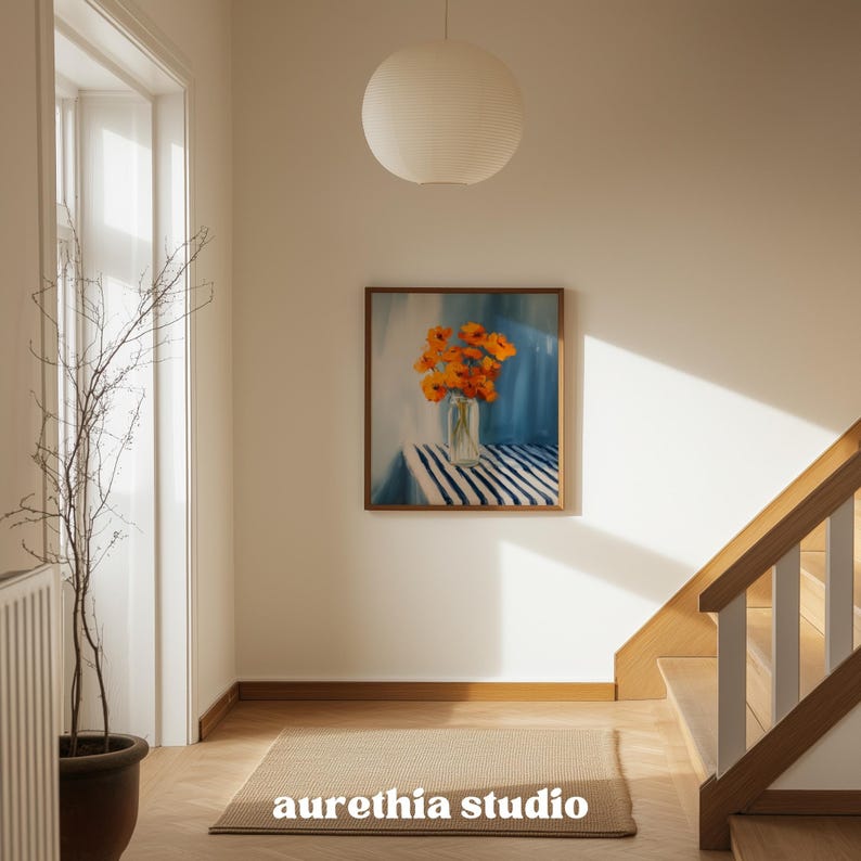 May include: A framed painting depicts orange flowers in a glass vase on a striped table. The artwork hangs on a white wall, above a rectangular rug. The text "aurethia studio" is at the bottom of the rug.