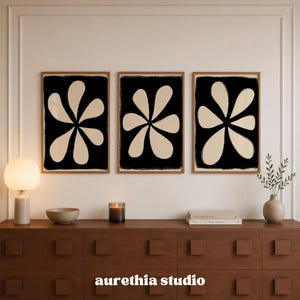 May include: Three rectangular wall art prints in light wood frames, each featuring a large, beige, six-petaled flower shape centered on a solid black background. The text "aurethia studio" is printed in white at the bottom center of the image.