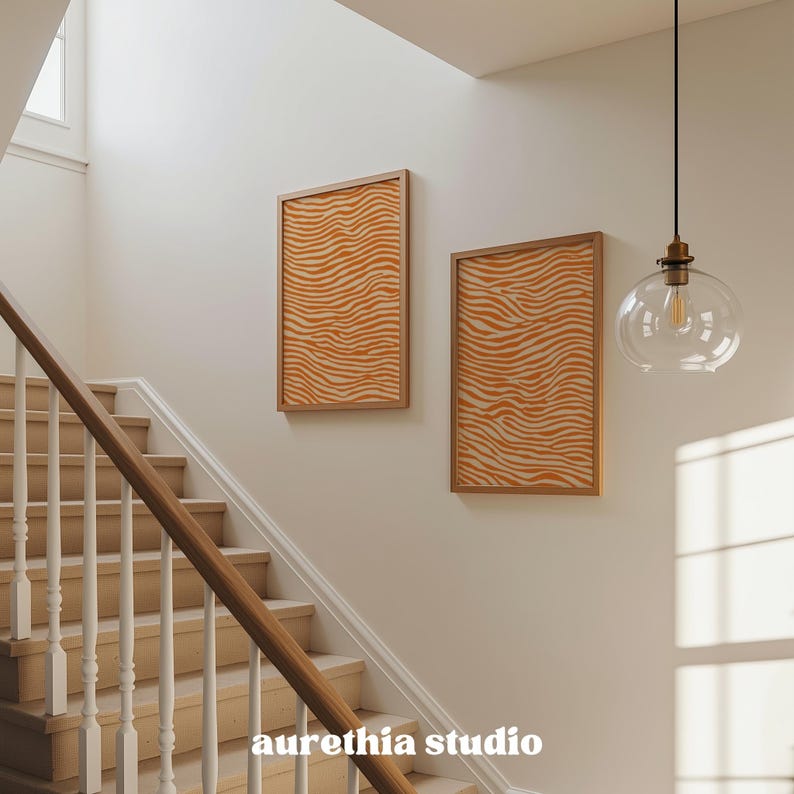 May include: Two framed art prints with an orange and cream zebra stripe pattern hang on a cream-colored wall. The prints are in wooden frames. A staircase with wooden steps and a wooden handrail is in the foreground. The text "aurethia studio" is at the bottom.