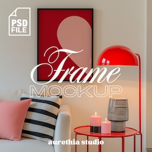 May include: A white couch with pink and black and white striped pillows sits in front of a white wall with a black framed picture of a red and pink abstract design. A red lamp with a white shade is on the right side of the image. A red side table with two candles and a black and white striped vase sits in front of the couch.