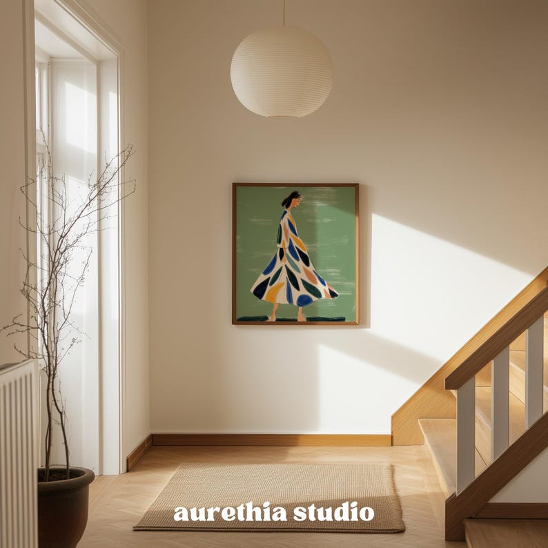 May include: A framed artwork featuring a figure in a flowing dress with colorful leaf-like patterns against a green background. The artwork hangs on a white wall in a well-lit hallway. A beige rug and a hanging paper lantern are also visible.