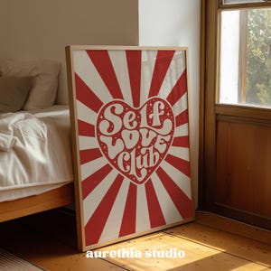 May include: A framed art print with a retro design. The artwork features a red and white sunburst pattern with a heart-shaped center. The words "Self Love Club" are written in a groovy font within the heart. The print is displayed on a wooden floor.