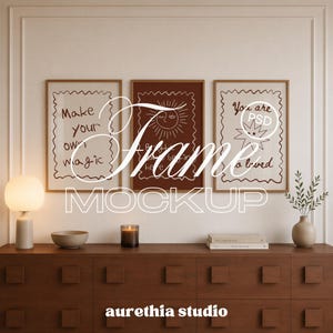 May include: Three rectangular wooden frames containing white and dark brown prints with line art of a sun and stars, featuring the text Make your own magic, You are loved, and other phrases, displayed on a dark wooden cabinet with a lamp and vase.