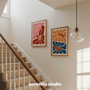 May include: Two framed floral art prints with a mid-century modern aesthetic. One print features pink and red flowers, the other orange and blue. The prints are displayed on a cream-colored wall above a wooden staircase. The text "aurethia studio" is at the bottom.