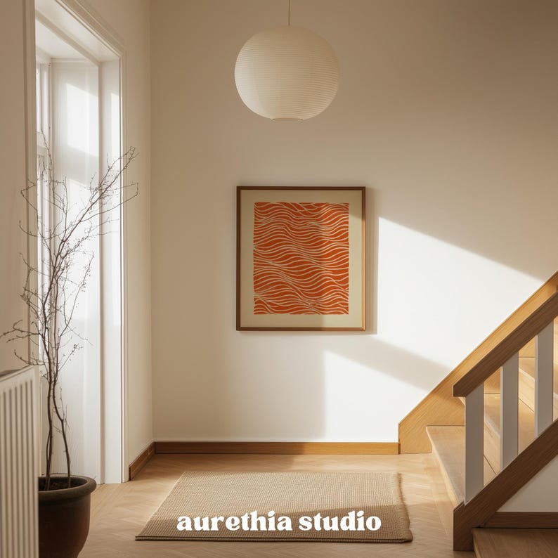 May include: A framed orange and white abstract art print hangs on a white wall in a well-lit hallway. The print features a wavy pattern. A neutral-toned rug and a potted plant are in the foreground. The text "aurethia studio" is at the bottom of the rug.