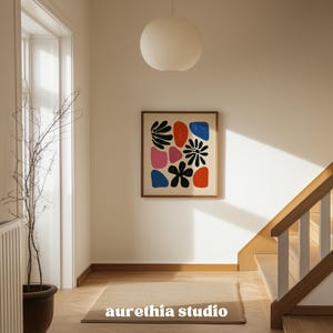 May include: A framed abstract art print featuring colorful shapes and floral designs in black, red, pink, and blue. The artwork hangs on a white wall in a well-lit hallway. The text "aurethia studio" is visible at the bottom of the image.