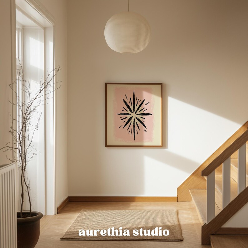 May include: A framed artwork with a black starburst design on a pink and yellow background hangs on a white wall. The artwork is in a room with a neutral color palette, a beige rug, and a wooden staircase. The text "aurethia studio" is at the bottom.