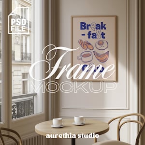Op de afbeelding: Een ingelijste poster-mockup met het woord "Breakfast" in blauw en illustraties van ontbijtproducten. De poster staat op een crèmekleurige muur. Een kleine ronde tafel met twee witte kopjes en schotels staat voor een raam.