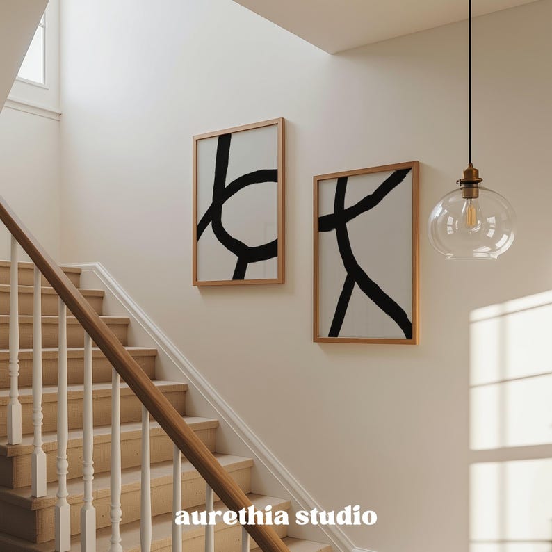 May include: Two framed abstract art prints with black brushstrokes on a white background hang on a wall. The frames are a light wood color. A staircase with wooden steps and a white banister is in the foreground. A clear glass pendant light hangs from the ceiling.