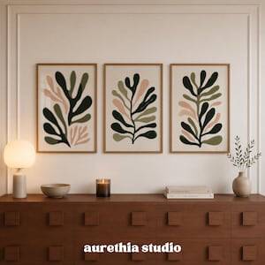May include: Three framed wall art prints featuring abstract botanical leaf shapes in shades of dark green, olive green, and soft pink on a light beige background. The prints are arranged horizontally in matching light wood frames on a wall.