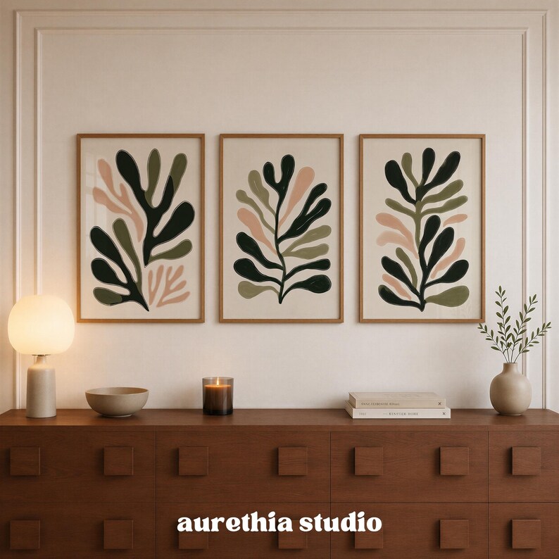 May include: Three framed wall art prints featuring abstract botanical leaf shapes in shades of dark green, olive green, and soft pink on a light beige background. The prints are arranged horizontally in matching light wood frames on a wall.