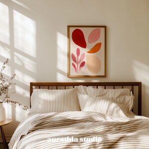 May include: A framed abstract art print with red, pink, and orange shapes hangs above a bed with striped bedding. The wooden bed frame and the neutral color palette create a calming bedroom scene. The art print features a botanical design.