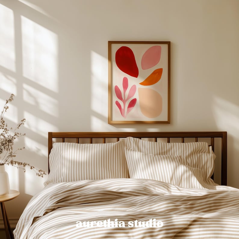 May include: A framed abstract art print with red, pink, and orange shapes hangs above a bed with striped bedding. The wooden bed frame and the neutral color palette create a calming bedroom scene. The art print features a botanical design.