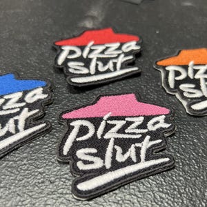 May include: Embroidered patches with the text "Pizza Slut" in white script. Each patch features a black background with a colored roof design in red, blue, pink, and orange. The patches are likely for clothing or accessories.