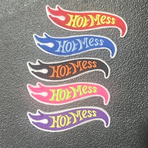 May include: Five embroidered patches in a flame design, each with the words "Hot Mess" in a stylized font. The patches are in red, blue, black, pink, and purple, with contrasting text colors of yellow, light blue, orange, green, and yellow.