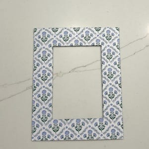 May include: A rectangular picture frame featuring a repeating pattern of small blue flowers with green stems and leaves on a white background. The frame has a flat, wide border surrounding a central rectangular opening for a photograph or artwork.
