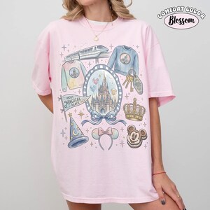 Puede incluir: Una camiseta rosa claro Comfort Colors con un dise&ntilde;o tem&aacute;tico de Disney. El dise&ntilde;o incluye un castillo, un tren, una corona, llaves y otros elementos m&aacute;gicos. Tambi&eacute;n se incluyen las palabras "Most Magical Place".