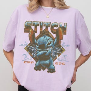 Peut inclure: T-shirt lilas avec un motif de Stitch de Lilo & Stitch. Le motif comprend le mot "STITCH" dans une police vieillie au-dessus de Stitch, avec "626" en dessous. Stitch est bleu avec de grandes oreilles et les bras crois&eacute;s.