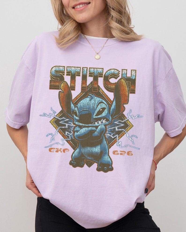Peut inclure: T-shirt lilas avec un motif de Stitch de Lilo & Stitch. Le motif comprend le mot "STITCH" dans une police vieillie au-dessus de Stitch, avec "626" en dessous. Stitch est bleu avec de grandes oreilles et les bras crois&eacute;s.