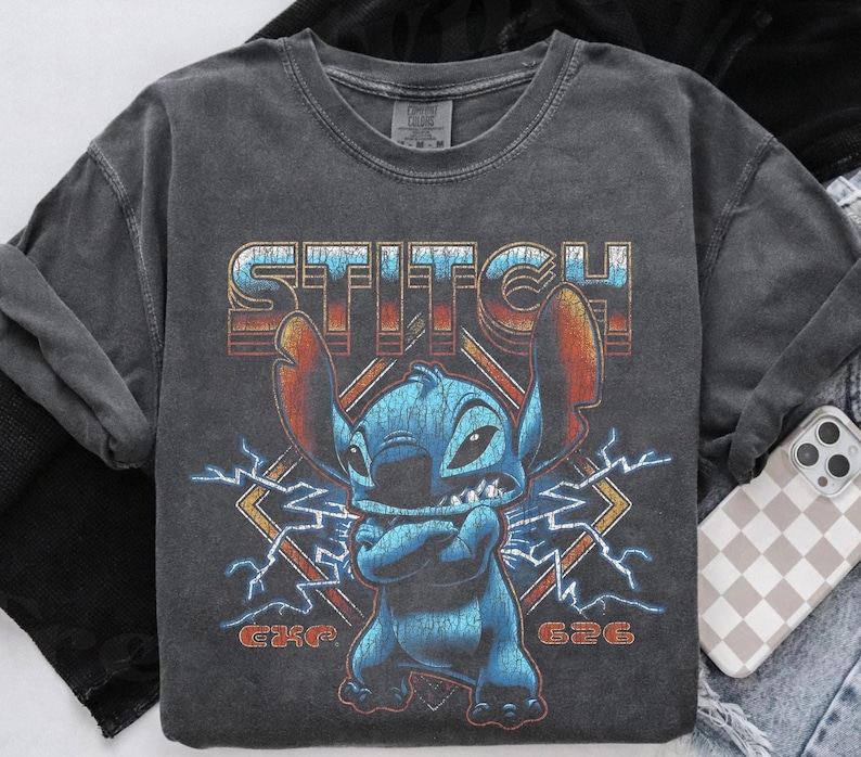 Peut inclure: T-shirt &agrave; manches longues gris fonc&eacute; avec un motif vieilli de Stitch, le mot "STITCH" au-dessus et "626" en dessous. Le personnage bleu Stitch a les bras crois&eacute;s et est entour&eacute; d'&eacute;clairs. Le t-shirt a un aspect vintage et us&eacute;.