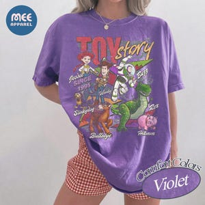 Pu&ograve; includere: Una t-shirt viola a maniche corte con una stampa grafica dei personaggi di Toy Story, tra cui Jessie, Woody, Buzz Lightyear, Slinky Dog, Bullseye, Rex e Hamm. Il testo riporta Toy Story, Since 1995, Jessie, Woody, Buzz, Slinky Dog, Bullseye, Rex e Hamm.