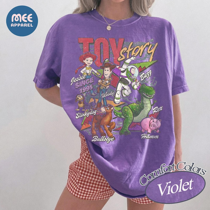 Pu&ograve; includere: Una t-shirt viola a maniche corte con una stampa grafica dei personaggi di Toy Story, tra cui Jessie, Woody, Buzz Lightyear, Slinky Dog, Bullseye, Rex e Hamm. Il testo riporta Toy Story, Since 1995, Jessie, Woody, Buzz, Slinky Dog, Bullseye, Rex e Hamm.