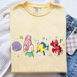 May include: A pale yellow t-shirt featuring a graphic print of a purple seashell, a character with long pink hair, a yellow and blue fish, and a red crab. The design includes small yellow stars and blue paint splatters across the center chest.
