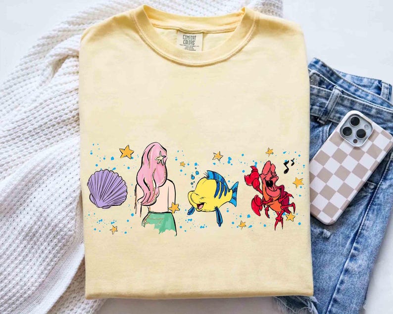 May include: A pale yellow t-shirt featuring a graphic print of a purple seashell, a character with long pink hair, a yellow and blue fish, and a red crab. The design includes small yellow stars and blue paint splatters across the center chest.