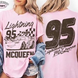 May include: A light pink t-shirt featuring a graphic of Lightning McQueen from the Cars movie. The front design includes the text "Lightning 95 McQueen" with an image of the race car. The back of the shirt displays a large "95" and "Lightning McQueen".