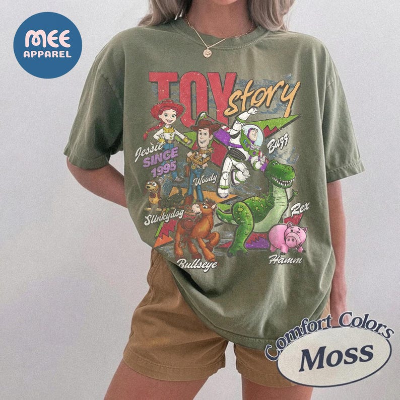 Pu&ograve; includere: Una t-shirt a maniche corte color verde oliva con una stampa grafica in stile vintage di Toy Story. Il design include i personaggi Jessie, Woody, Buzz Lightyear, Slinky Dog, Rex e Hamm con i loro nomi e il testo "SINCE 1995" in un carattere usurato.