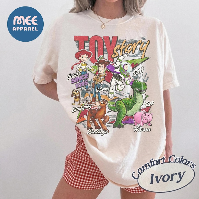 Pu&ograve; includere: Una t-shirt color avorio con una stampa grafica colorata dei personaggi di Toy Story, tra cui Woody, Buzz Lightyear, Jessie, Slinky Dog, Bullseye, Rex e Hamm. Il testo recita Toy Story, Jessie Since 1995, Woody, Buzz, Slinky Dog, Bullseye, Rex e Hamm.