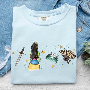 May include: A light blue t-shirt with a graphic print featuring a sword, a stylized figure with long black hair, a white flower hair comb, and a folding fan, accented by small yellow stars and green paint splatters. The neck label reads Comfort Colors.