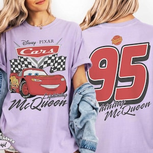 Pu&ograve; includere: Una maglietta lilla con Lightning McQueen di Cars di Disney Pixar. La grafica frontale include il logo Cars, una bandiera a scacchi e Lightning McQueen. Il retro mostra un grande "95" e la scritta "Lightning McQueen".