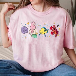 May include: A light pink t-shirt featuring a graphic print of a purple seashell, a mermaid with long pink hair, a yellow and blue fish, and a red crab, all surrounded by small yellow stars and blue paint splatter accents on the front.