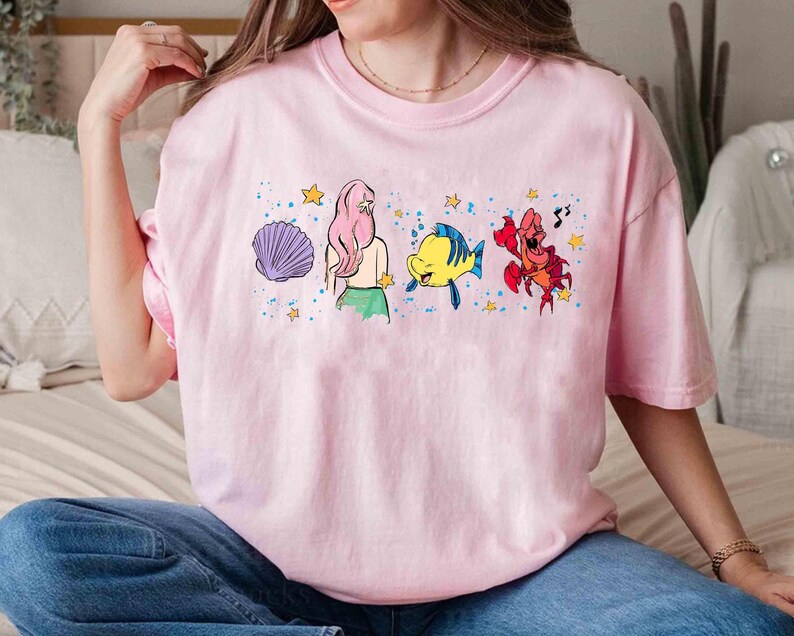 May include: A light pink t-shirt featuring a graphic print of a purple seashell, a mermaid with long pink hair, a yellow and blue fish, and a red crab, all surrounded by small yellow stars and blue paint splatter accents on the front.