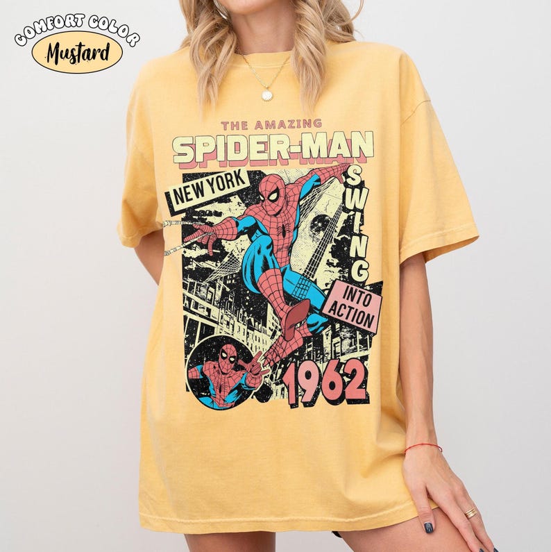 Puede incluir: Camiseta de color mostaza con un gr&aacute;fico vintage de Spider-Man. El dise&ntilde;o incluye el texto "THE AMAZING SPIDER-MAN", "NEW YORK" y "1962". El gr&aacute;fico muestra a Spider-Man en acci&oacute;n, con las palabras "SWING INTO ACTION".