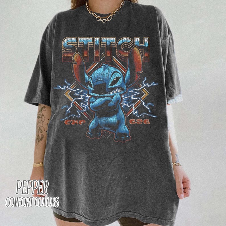 Peut inclure: T-shirt gris fonc&eacute; avec un personnage de dessin anim&eacute; bleu et le mot "STITCH" dans une police r&eacute;tro. Le motif comprend des &eacute;clairs et le texte "G24G" et "G36G".