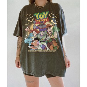 May include: An olive green oversized t-shirt featuring the Toy Story logo and characters. The graphic includes Buzz Lightyear, Woody, and other characters from the animated film. The shirt has a vintage, worn look.