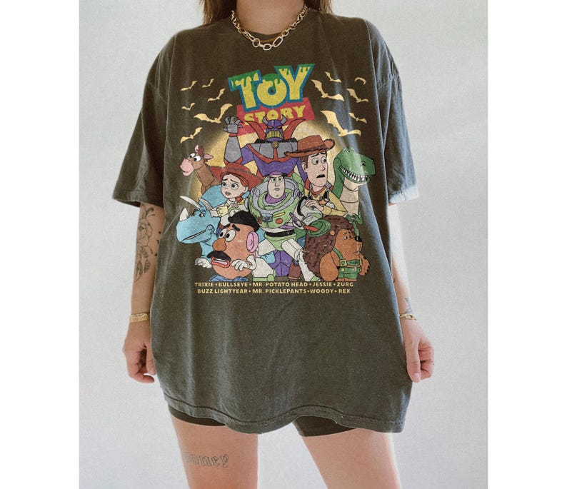 May include: An olive green oversized t-shirt featuring the Toy Story logo and characters. The graphic includes Buzz Lightyear, Woody, and other characters from the animated film. The shirt has a vintage, worn look.