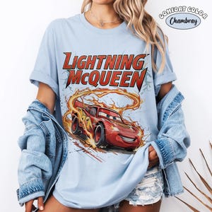Op de afbeelding: Lichtblauw T-shirt met een afbeelding van Lightning McQueen uit de film Cars. Het ontwerp bevat de tekst "LIGHTNING McQUEEN" in rood en geel, met het personage in beweging met vlammen.
