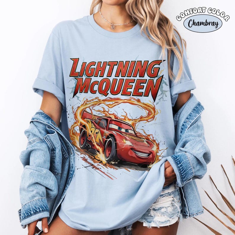 Op de afbeelding: Lichtblauw T-shirt met een afbeelding van Lightning McQueen uit de film Cars. Het ontwerp bevat de tekst "LIGHTNING McQUEEN" in rood en geel, met het personage in beweging met vlammen.