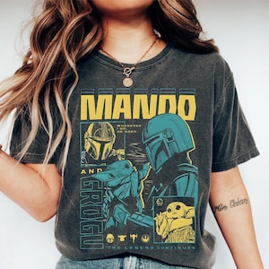 Pode incluir: Camiseta cinza escuro com a palavra "MANDO" em letras amarelas. A camiseta apresenta ilustra&ccedil;&otilde;es do Mandaloriano e Grogu em verde-azulado e amarelo. O texto "WHEREVER HE GOES" e "THE LEGEND CONTINUES" tamb&eacute;m est&atilde;o presentes.
