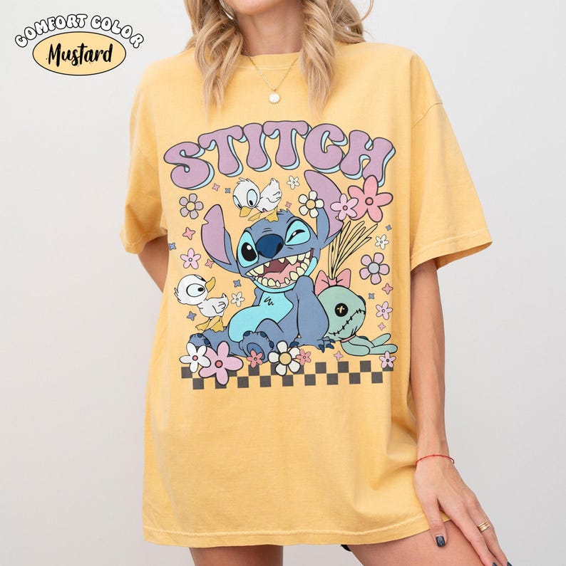 May include: Mustard-coloured t-shirt with the word "Stitch" in a retro font, and a cartoon Stitch character, flowers, and other characters. The shirt has a relaxed fit and short sleeves. The text "Comfort Color" is in the upper left corner.