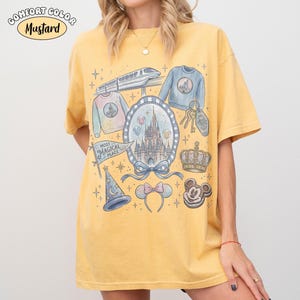 Puede incluir: Camiseta de color mostaza con un gr&aacute;fico que presenta un castillo, un tren, una corona, Mickey Mouse y otros elementos tem&aacute;ticos de Disney. La camiseta tiene las palabras "Most Magical Place" en una pancarta y "Comfort Color" en la parte superior izquierda.