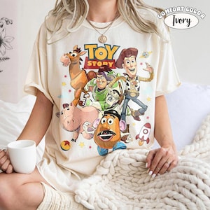 May include: Ivory-colored t-shirt featuring a graphic of characters from the animated film "Toy Story." The design includes Woody, Buzz Lightyear, Mr. Potato Head, and others. The shirt has the words "Toy Story" in a yellow and brown font.