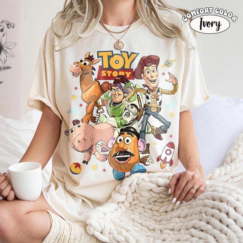 May include: Ivory-colored t-shirt featuring a graphic of characters from the animated film "Toy Story." The design includes Woody, Buzz Lightyear, Mr. Potato Head, and others. The shirt has the words "Toy Story" in a yellow and brown font.